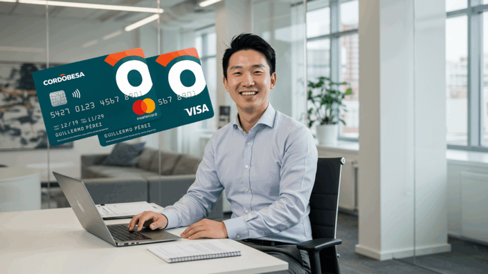 Hombre joven sonriente sentado frente a una laptop en una oficina moderna. A la izquierda, se visualizan dos tarjetas Cordobesa, una Mastercard y una Visa, con diseño contemporáneo en tonos azulados.