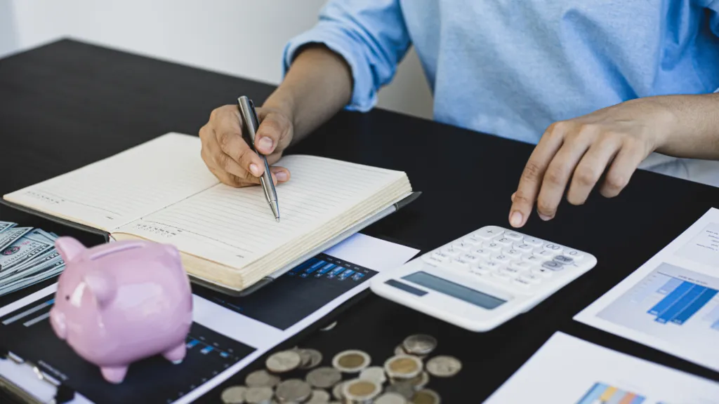 A person with a piggy bank, coins, and a calculator on the table taking notes about saving-money.