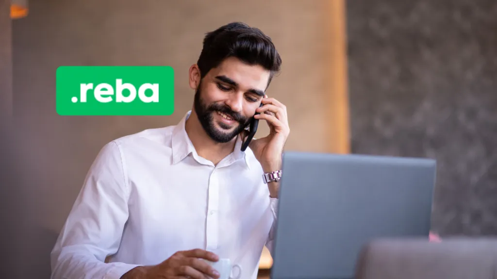 Hombre con barba, vistiendo una camisa blanca, sonriendo mientras habla por teléfono y sostiene una taza, frente a una computadora portátil.