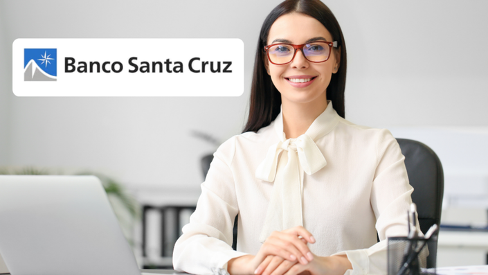 Mujer sonriendo, sentada en su escritorio, con las manos sobre la mesa, y con el logo de "Banco Santa Cruz" en la esquina superior izquierda.