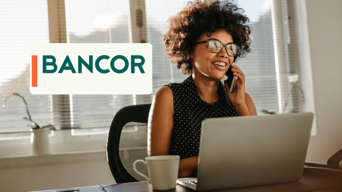 Mujer sonriente hablando por teléfono mientras trabaja con una computadora portátil en una oficina iluminada, con el logo de BANCOR. Imagen relacionada con préstamos Bancor.