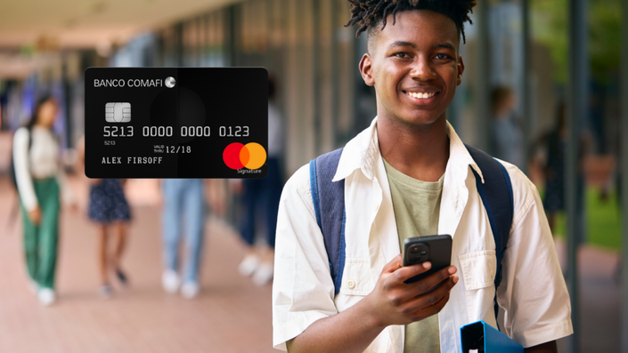 Un joven sonriente, sosteniendo un teléfono móvil mientras camina por un pasillo de su universidad. En la esquina superior derecha, se muestra una tarjeta de crédito de Banco Comafi con un diseño negro y el logo de Mastercard.