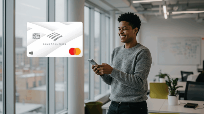 Joven sonriente utilizando un teléfono móvil mientras sostiene una tarjeta de crédito Bank of America Mastercard. La imagen refleja un ambiente dinámico y tecnológico.