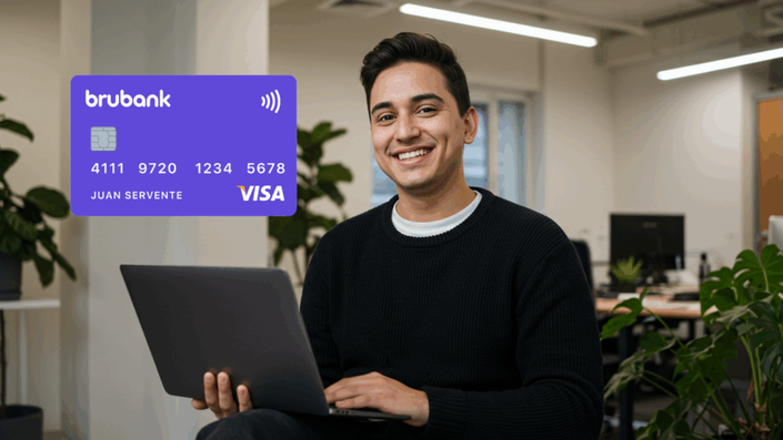 Un joven sorrindo enquanto usa um laptop, com uma tarjeta de crédito Brubank em destaque. A tarjeta é de cor roxa com o logo de Visa.