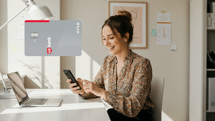 Mujer sonriente usando su teléfono móvil mientras sostiene una tarjeta de crédito US Bank Visa. La imagen refleja un ambiente relajado y tecnológico.