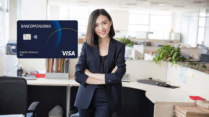 Mujer con traje formal oscuro, brazos cruzados, sonriendo en una oficina, junto a una tarjeta de crédito Visa del Banco Patagonia.