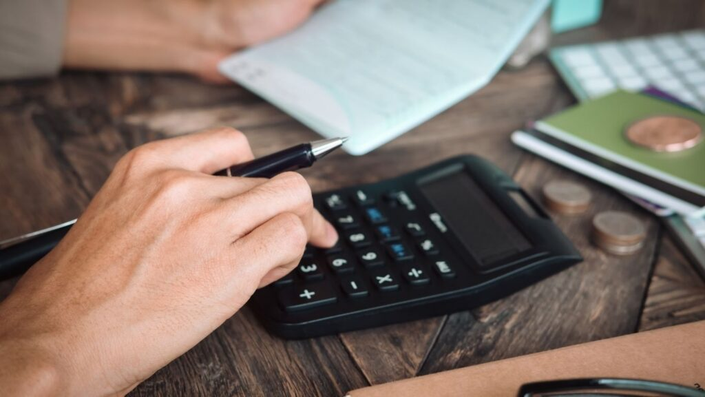 Persona usando calculadora científica mientras sostiene una lapicera, con documentos financieros desparramados sobre mesa de madera rústica, monedas apiladas al costado y cuaderno verde al fondo. Imagen representativa del proceso para consolidar deudas y organizar mejor tu planificación financiera