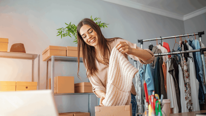 Joven emprendedora sonriendo mientras empaca con cuidado una prenda de lana en una caja para envío, en su espacio de trabajo con estanterías llenas de paquetes y un perchero con ropa, ilustrando la gestión de un e-commerce de moda.
