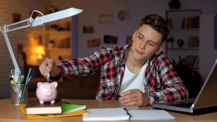 Joven ahorrando dinero en una alcancía mientras estudia en su escritorio. La educación financiera para niños y jóvenes ayuda a desarrollar hábitos de ahorro desde temprana edad.