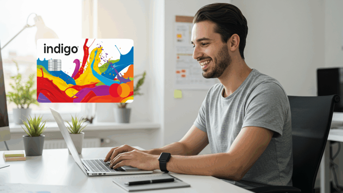 Hombre joven sonriente usando una laptop en una oficina iluminada. A la izquierda, se muestra una tarjeta de crédito Indigo® Mastercard® con un diseño colorido de salpicaduras de pintura.