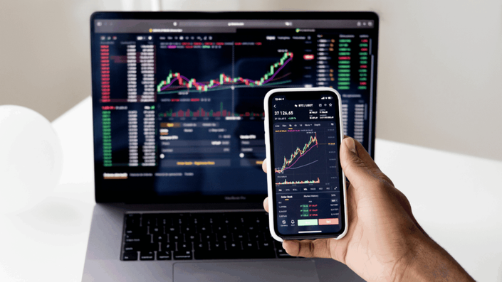 Una mano sosteniendo un smartphone con gráficos de trading de criptomonedas, mientras una laptop abierta en el fondo muestra gráficos similares, representando la dinámica y la tecnología del mercado cambiario en Argentina.