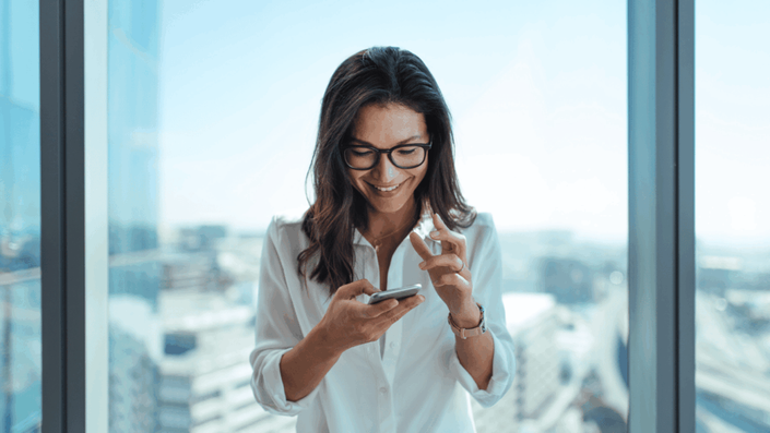 Mujer joven sonriente consultando su inversión de plazo fijo en Argentina desde su celular, en una oficina moderna con vista panorámica.