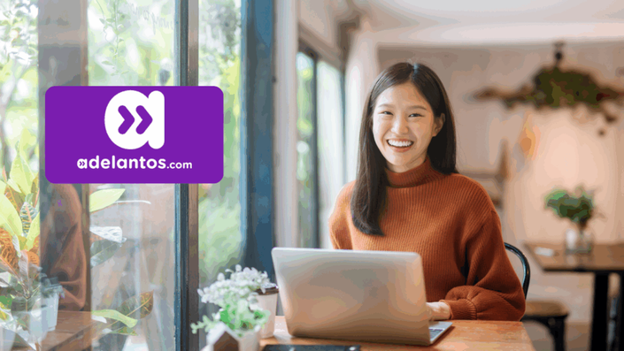 Mujer joven sonriente con suéter naranja trabajando en una laptop junto a una ventana con plantas. A la izquierda, se muestra el logo de adelantos.com en color violeta.